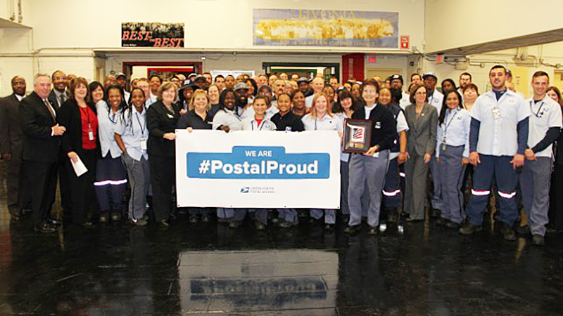 PMG Megan Brennan, fifth from left on the front row, and Livonia, MI, Post Office employees display a #PostalProud banner.