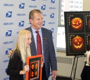 Chief Financial Officer Joe Corbett (right) and St. Stephen’s Catholic School student Samantha Jenson (left) pose for a picture with a pane of Jack-o’-lantern stamps.