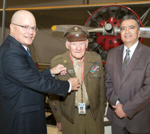 Williams presents a pin to Wilbur Richardson, a World War II veteran and docent at the Planes of Fame Air Museum, with help from Santa Ana District Manager Eduardo Ruiz.