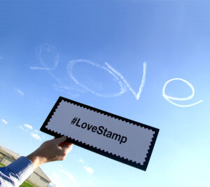 The word “Love” appears in the skies above Chino while Pacific Area Writer/Editor John Hyatt reminds everyone to use the hashtag #LoveStamp to discuss the stamps on social media.