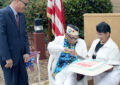 Veteran dressed in white sits and cuts cake with help from two others