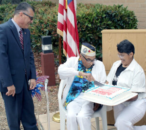 Veteran dressed in white sits and cuts cake with help from two others