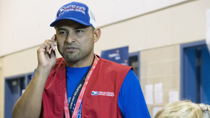 Man talks on phone with blue postal hat and red vest