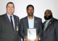 Two men flank a third who holds a plaque