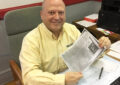 Smiling man sits at desk, holding newspaper clipping