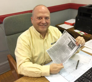 Smiling man sits at desk, holding newspaper clipping