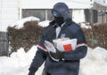 Letter carrier in heavy winter gear walks through snowy neighborhood