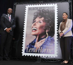 Two people stand on stage next to oversized Lena Horne stamp poster