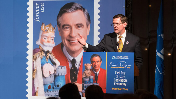 Man stands at podium near Mister Rogers stamp poster