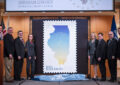 People stand on stage next to stamp poster