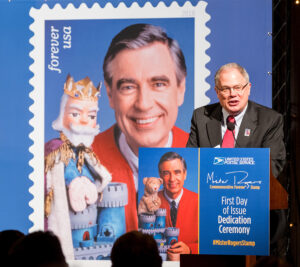 Man speaks at podium near Mister Rogers stamp poster