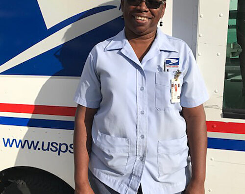 Smiling woman in postal uniform stands next to delivery vehicle