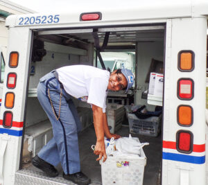 Smiling postal worker stands in back of delivery vehicle, sorting bags of food