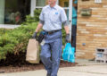 Smiling postal worker carries bags of food down home's driveway