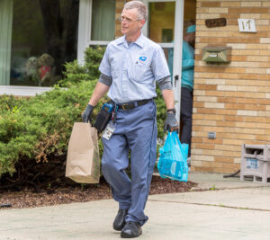 Smiling postal worker carries bags of food down home's driveway