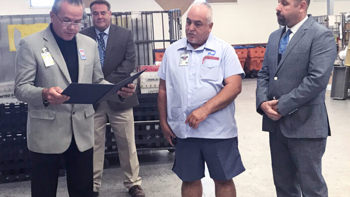 Man in business suit presents certificate to two postal workers as fourth man watches