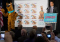 Man in suit speaks at podium with costumed character nearby