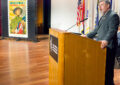 Man speaks at podium with stamp poster in background