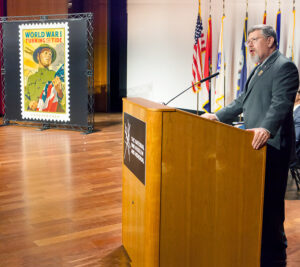 Man speaks at podium with stamp poster in background