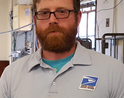 Man wearing USPS uniform stands in postal workroom