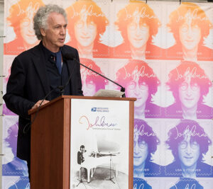 Man stands at podium near John Lennon stamp poster