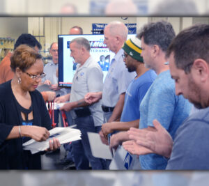 Woman hands envelopes to men who are lined up in postal workroom