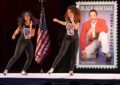 Two women dance on stage in front of Gregory Hines stamp poster