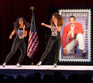 Two women dance on stage in front of Gregory Hines stamp poster