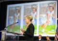 Woman speaks at podium near bank of video screens displaying Little Mo stamp image