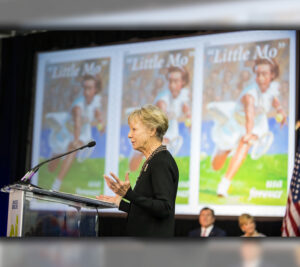 Woman speaks at podium near bank of video screens displaying Little Mo stamp image