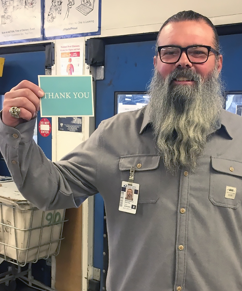 Smiling man stands in postal workroom, holding thank-you card