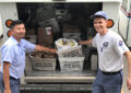 Two smiling postal workers stand near delivery vehicle filled with bags of food