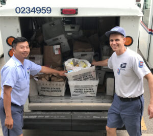 Two smiling postal workers stand near delivery vehicle filled with bags of food