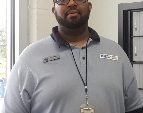 Man in USPS uniform stands inside Post Office lobby