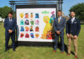 Three smiling men stand near oversized stamp artwork