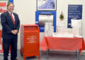 Smiling Postmaster stands inside Post Office lobby