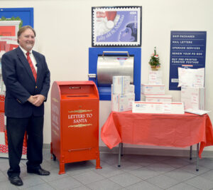 Smiling Postmaster stands inside Post Office lobby