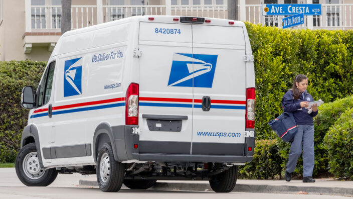 Postal employee walks pass a parked USPS vehicle.