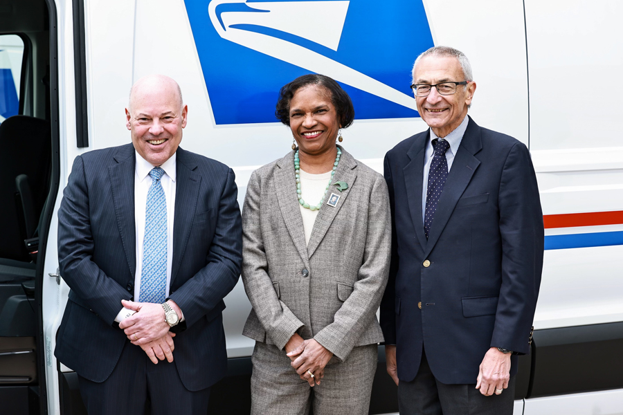 Three government officials stand near a USPS delivery vehicle