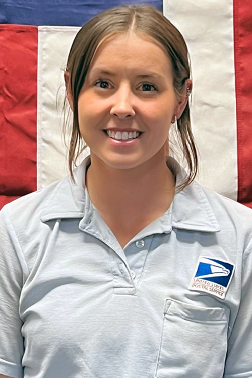 Smiling woman wearing postal uniform shirt stands in front of U.S. flag