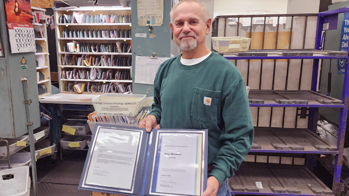 Smiling man displays certificate of recognition while standing in post office workroom