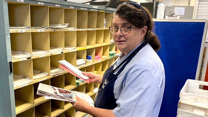 A woman in a postal uniform sorts mail at a cubby