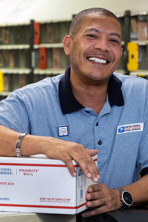 A smiling postal worker leans on a Priority Mail box on a retail countertop