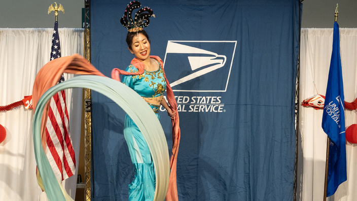 Asian woman performs dance on stage in front of curtain bearing USPS logo