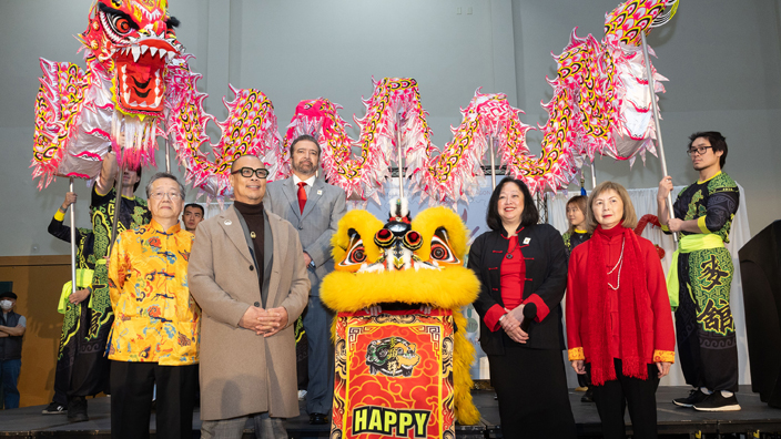 A group of people stand on a stage surrounded by festive Lunar New Year decor and a someone wearing a large dragon costume