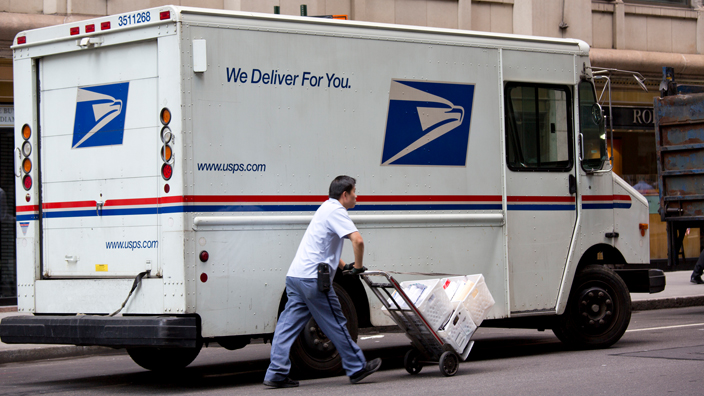Uniformed postal worker rolls cart loaded with packages near delivery truck