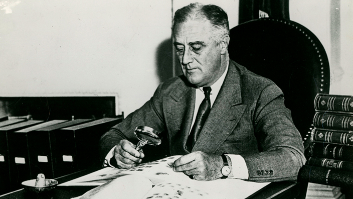 Black and white image of Franklin Roosevelt seated at a desk, using a magnifying glass to examine stamp album
