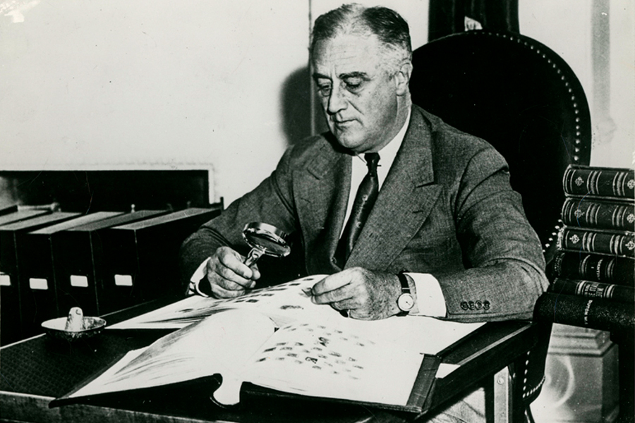Black and white image of Franklin Roosevelt seated at a desk, using a magnifying glass to examine stamp album