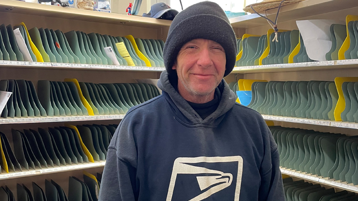 Man wearing a postal hoodie stands in front of mail cubby