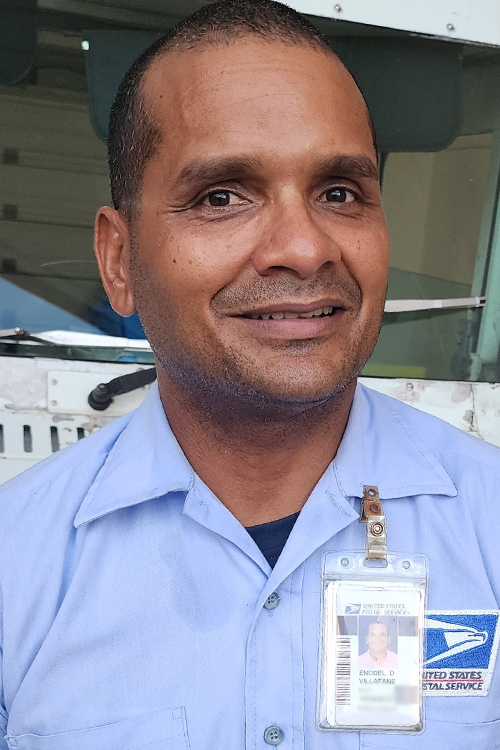 A smiling man in a postal uniform stands near a USPS delivery truck